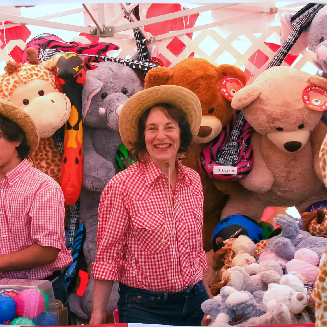 A smiling carnival worker in a straw hat stands in front of a prize booth filled with large stuffed animals at a corporate event in Los Angeles.